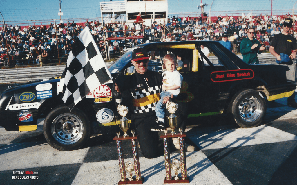 Race car driver celebrating victory with trophies and checkered flag.